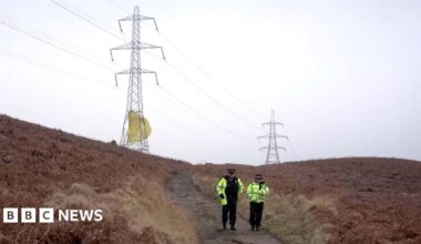 A male and a female police officer walk down a remote dirt track, which is flanked on both side by gorse. On the horizon are two large electricity pylons, there is yellow-coloured material that is wrapped around the bottom of the nearest pylon that appears to be billowing in the wind