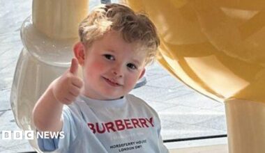 Peter Maughan is standing outside on a summer's day wearing a grey T-shirt with the Burberry logo in red capital letters. He is making a thumbs-up sign with his head tilted and he is smiling at the camera looking happy. He has short, blond hair and is face is flushed.