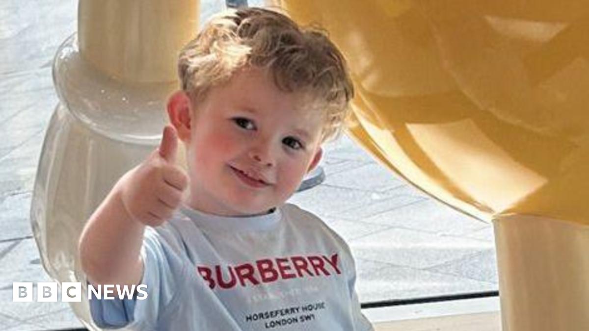 Peter Maughan is standing outside on a summer's day wearing a grey T-shirt with the Burberry logo in red capital letters. He is making a thumbs-up sign with his head tilted and he is smiling at the camera looking happy. He has short, blond hair and is face is flushed.