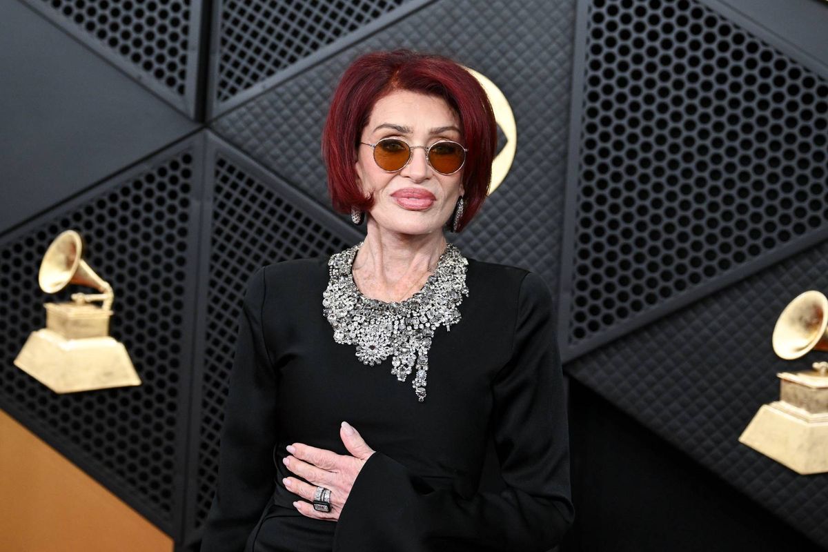 An individual with red hair is standing in front of a distinctive wall adorned with golden gramophone awards, dressed in a black long-sleeved outfit and sunglasses.