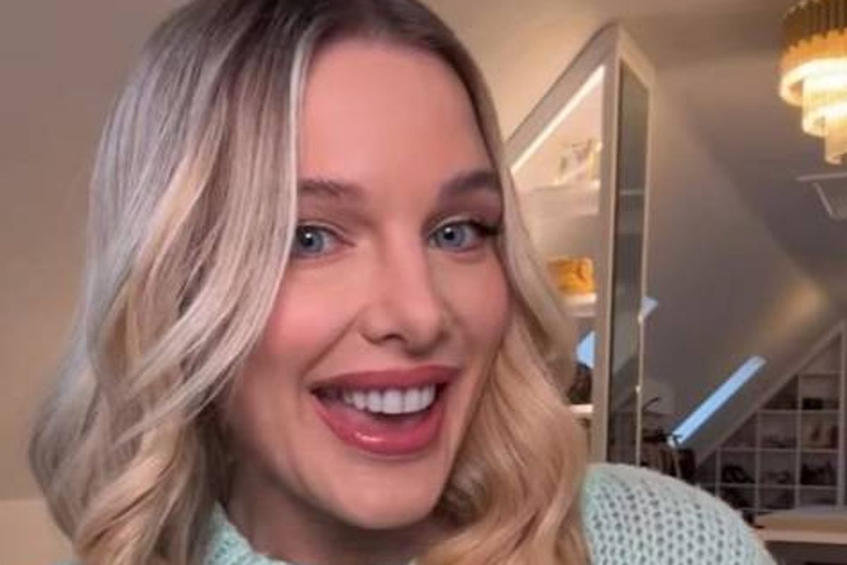 An individual with long, light-colored hair and a radiant smile, positioned in an interior setting with ambient lighting.