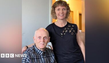 An elderly man in a buttoned up shirt sits next to a younger woman with fair curly hair who stands at his side.