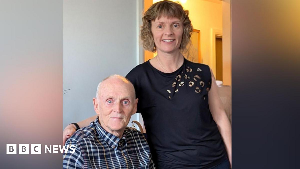 An elderly man in a buttoned up shirt sits next to a younger woman with fair curly hair who stands at his side.
