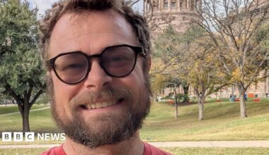 Close up of a man's face with beard moustache and glasses. He is standing outdoors against a backdrop of trees and grass. He is smiling.