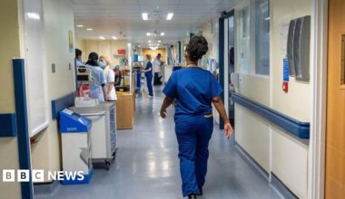 A nurse wears blue scrubs as she walks down a hospital corridor, there are other nurses in the background doing different tasks.