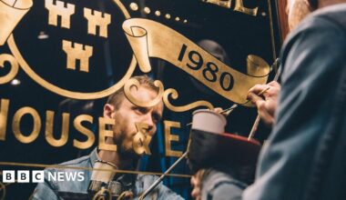 The outside of the Mean Eyed Cat, which is a small pub with a green exterior and pink sign. There are hanging baskets outside the shop, with large planters running along the windows. There is a sports shop next door and a row of terraced brick houses to the right.