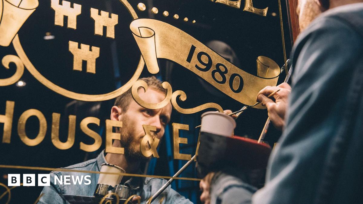 The outside of the Mean Eyed Cat, which is a small pub with a green exterior and pink sign. There are hanging baskets outside the shop, with large planters running along the windows. There is a sports shop next door and a row of terraced brick houses to the right.