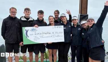 A group of people stood next to a lake. They are holding a large novelty cheque and smiling.