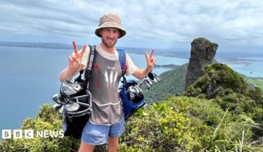 Dougie Haynes is doing a V peace symbol with both hands, whilst carrying two full golf bags on either shoulder. He is standing in front of a point where ocean meets land, with land visible on the other side of the water.