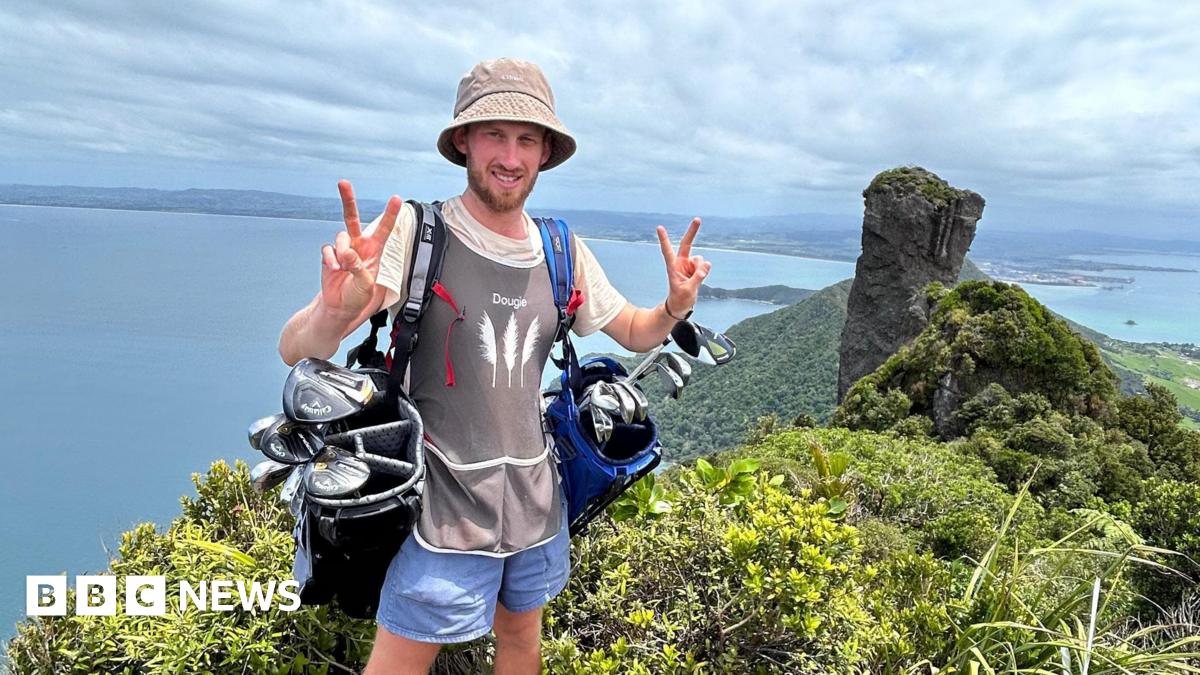 Dougie Haynes is doing a V peace symbol with both hands, whilst carrying two full golf bags on either shoulder. He is standing in front of a point where ocean meets land, with land visible on the other side of the water.