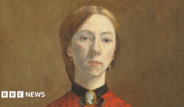 Oil painting of Gwen John. She is a white woman in her twenties. She has light red-brown hair pulled back into a low knot or bun at the back of her head. She is wearing an orange-red blouse with a check pattern on it. at her throat she is wearing a cameo broach with a black ribbon or collar around her throat. The clothing is consistent with the early 1900s. A black shawl is draped over her lower right arm.