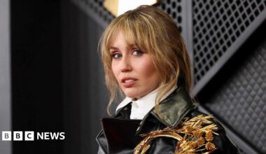 Miley Cyrus looks to the side of the camera wearing a black leather jacket and white shirt. She has brown/blonde hair with a pony-tail hairstyle.