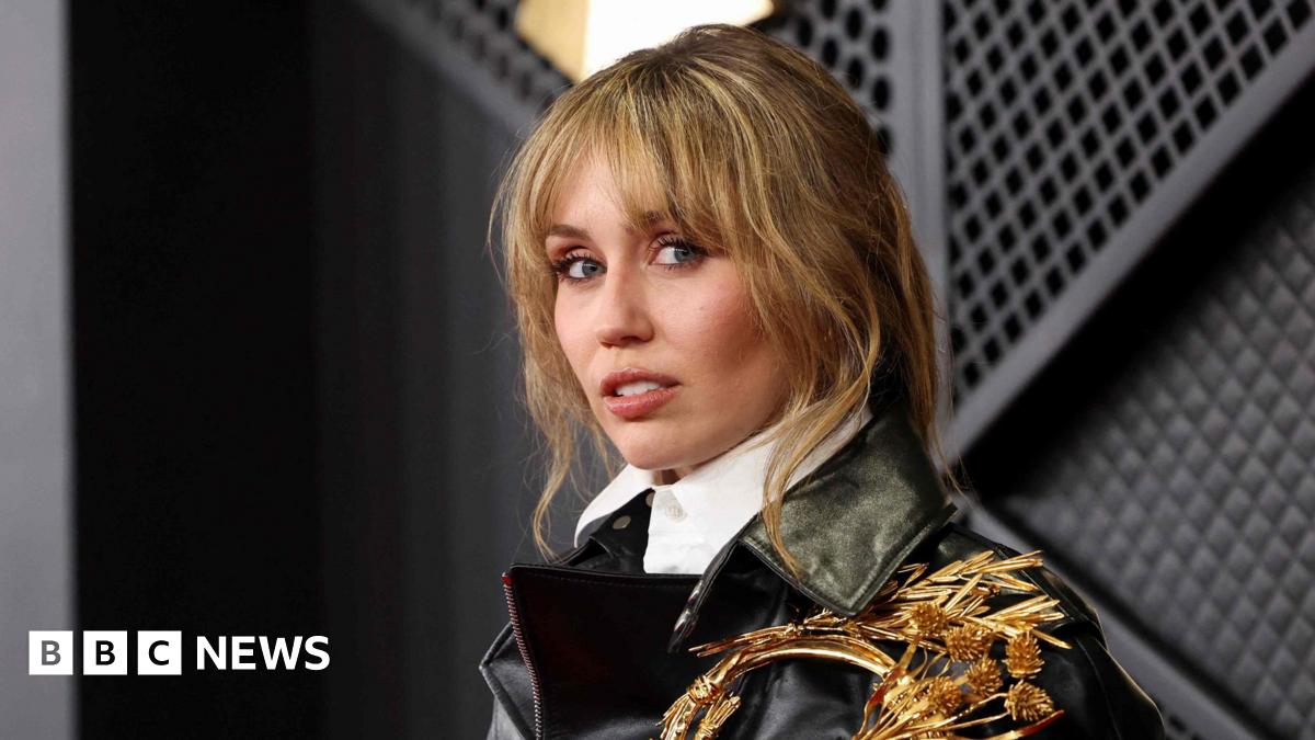 Miley Cyrus looks to the side of the camera wearing a black leather jacket and white shirt. She has brown/blonde hair with a pony-tail hairstyle.