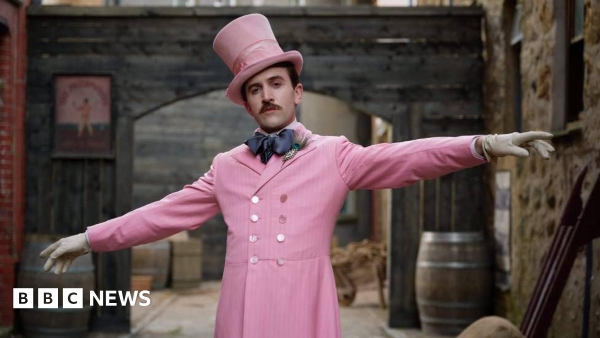 Callum Scott Howells dressed in one of Henry Paget's outfits. It is a pink buttoned up suit jacket paired with a large navy blue bow tie,. He is also wearing a pink top hat and has white gloves on his outstretched hands.