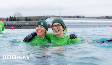 Two women in water wearing green swim rash vests and green woolly hats. They are both smiling and have their arms around each other.