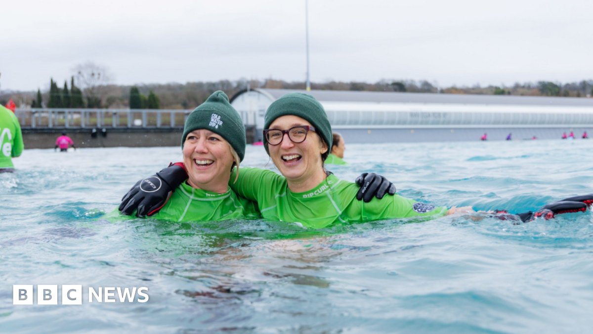 Two women in water wearing green swim rash vests and green woolly hats. They are both smiling and have their arms around each other.