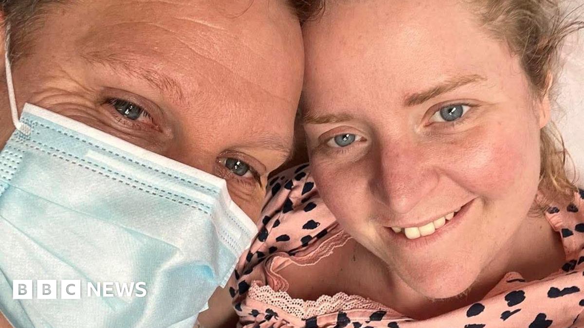 Melinda Tidy and her husband smiling with their heads leaning together. He is wearing a medical mask. She has long blonde hair piled in a bun on her head and blue eyes. He has dark hair and blue eyes.