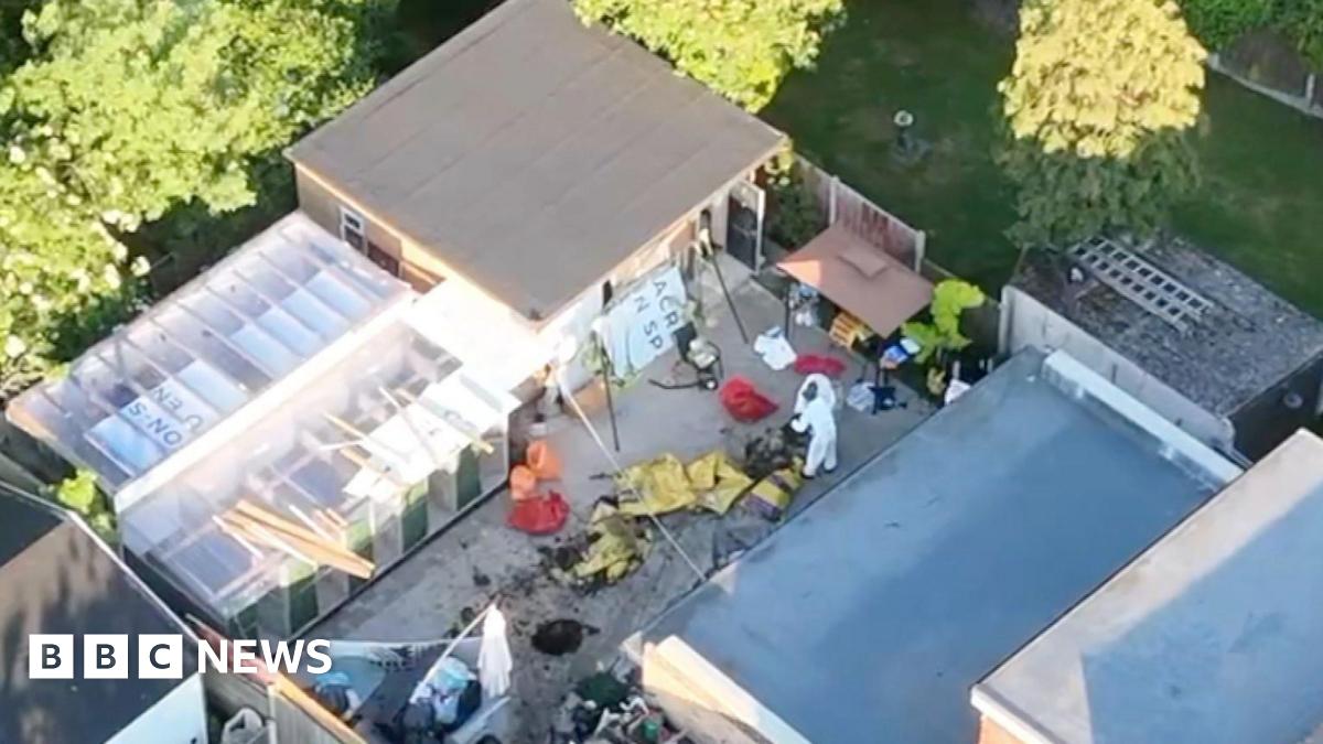 A drone image of the backyard at Save A Paw centre. Forensic officers wearing white jumpsuits are examining the scene, which is messy with various bags, mud and yellow sheets laid across it. There is a large grey outbuilding with a flat roof. Next to it is a smaller building with transparent roof.