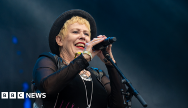 A woman with short blonde hair, a black top, black hat and long black gloves holding a black microphone in front of a dark background