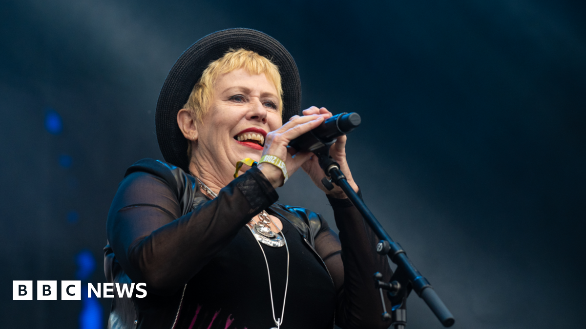 A woman with short blonde hair, a black top, black hat and long black gloves holding a black microphone in front of a dark background