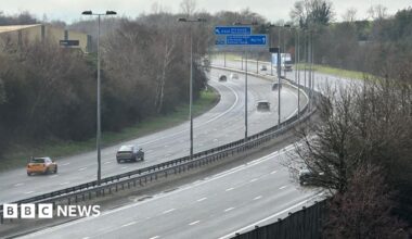 A bend in the M6 toll with some blue signs above it. There is a central reservation and a number of cars and a lorry travelling on both sides of the road. There are trees on the sides of the road.