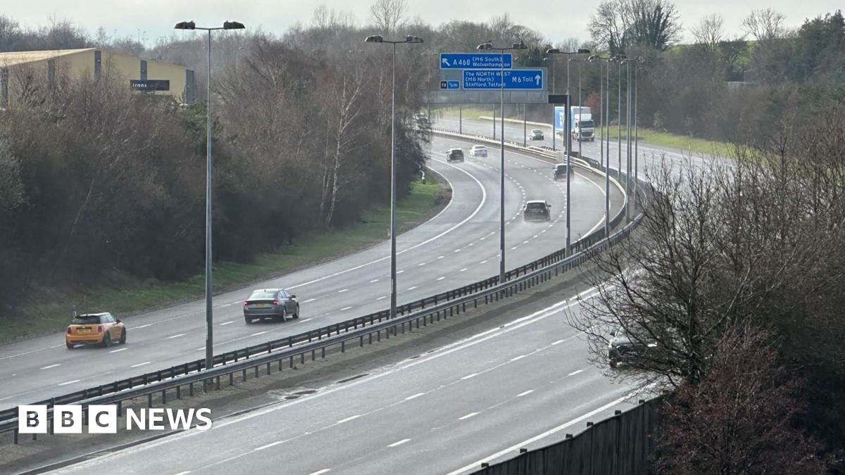 A bend in the M6 toll with some blue signs above it. There is a central reservation and a number of cars and a lorry travelling on both sides of the road. There are trees on the sides of the road.