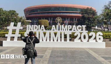 A man wearing a black pack takes a photo of a sign for the event, which says "Hashtag India AI Impact Summit 2026" in large white standalone letters. The sign sits in front of a round copper coloured building.