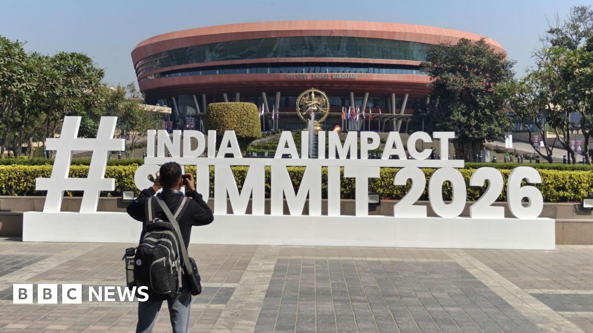 A man wearing a black pack takes a photo of a sign for the event, which says "Hashtag India AI Impact Summit 2026" in large white standalone letters. The sign sits in front of a round copper coloured building.