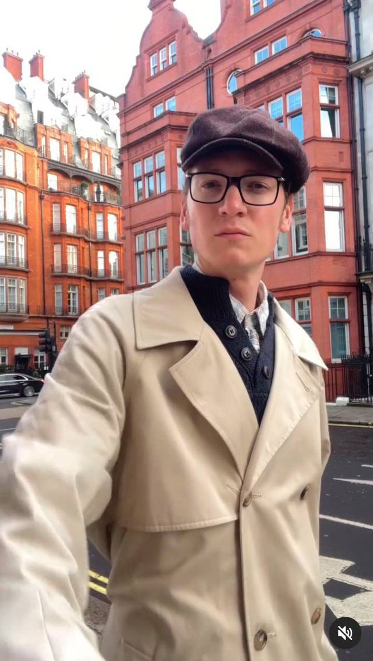 Photo of a bespectacled young man, wearing a dark brown flat cap, white shirt and blue jumper underneath a beige overcoat. Behind him are a few redbrick houses.