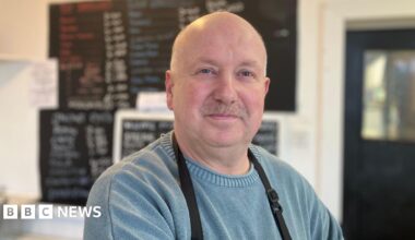 A man in a blue jumper at a sandwich shop. He wears an apron. Behind him a blackboard with food options is blurred.