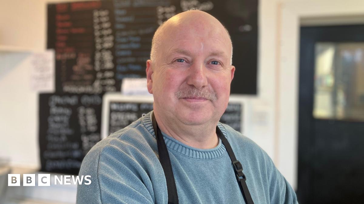 A man in a blue jumper at a sandwich shop. He wears an apron. Behind him a blackboard with food options is blurred.