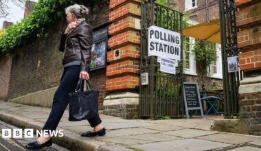 A woman holding a black bag walks away from a set of open metal gates, which has a white sign with 'polling station' written in black text on it.