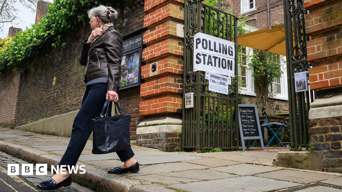 A woman holding a black bag walks away from a set of open metal gates, which has a white sign with 'polling station' written in black text on it.