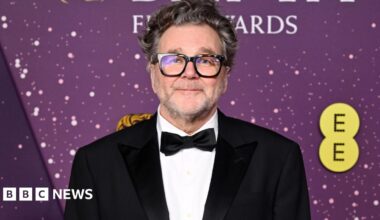 Kirk Jones. He has short grey hair and stubble. He is wearing black rimmed glasses, a white shirt, a black suit jacket and a black bowtie. He is looking directly at the camera and smiling.