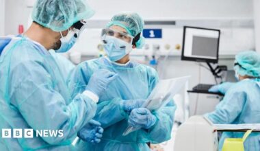 Two doctors talking wearing protective clothing. They are both wearing a blue gown and gloves with one holding a clipboard. There is another doctor looking at a monitor on the right.