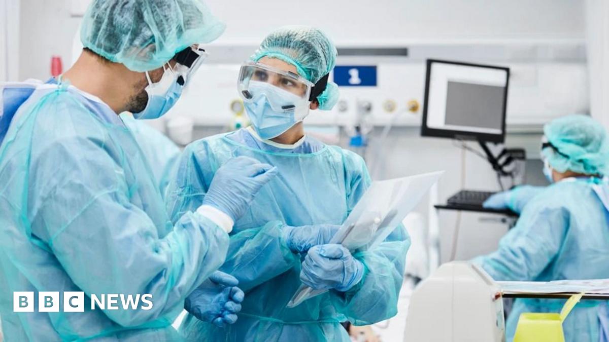 Two doctors talking wearing protective clothing. They are both wearing a blue gown and gloves with one holding a clipboard. There is another doctor looking at a monitor on the right.