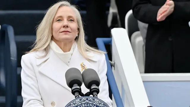 A woman with blonde hair, wearing a white suit and a gold broach stands at a lectern behind two microphones