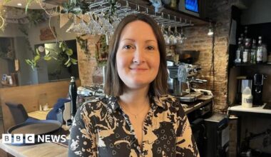 A woman in a patterned shirt stands smiling behind a cosy bar counter, with hanging glasses, bottles and a coffee machine in the background.