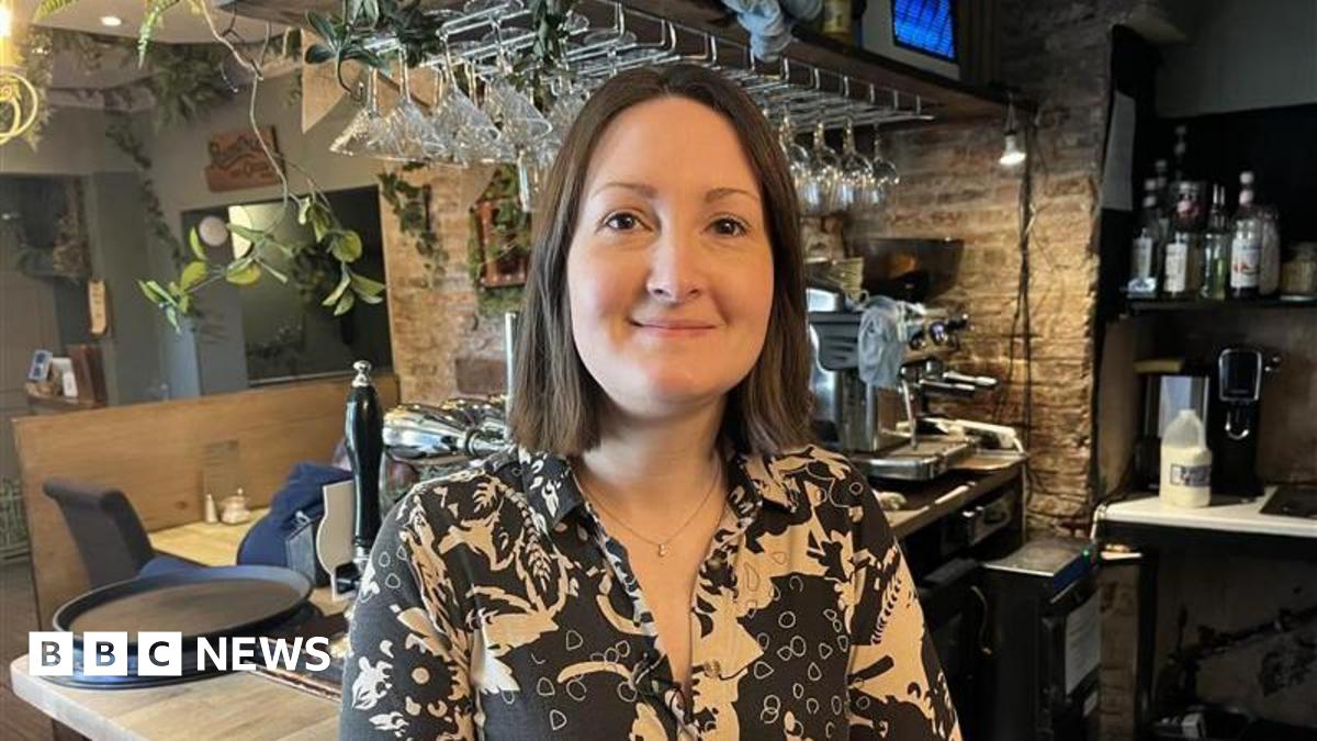 A woman in a patterned shirt stands smiling behind a cosy bar counter, with hanging glasses, bottles and a coffee machine in the background.