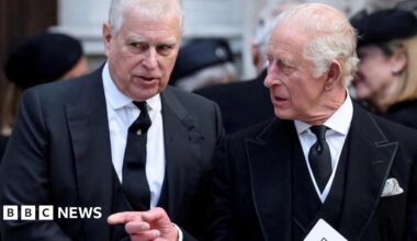Andrew and King Charles talking as they walk. Charles points across Andrew and looks to the side while Andrew looks ahead. They both wear black suits, white shirts and black ties