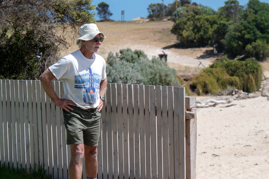 A man in a bucket hat, sunglasses, shorts and a tee shirt stands next to a broken back fence, with beach below it