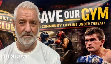 Steve Strong has white hair and a matching beard. He is wearing a white t-shirt and stands in front of a "Save Our Gym" banner which includes a photograph of two amateur boxers sparring, and a bloodied Ricky Hatton from one of his fights.