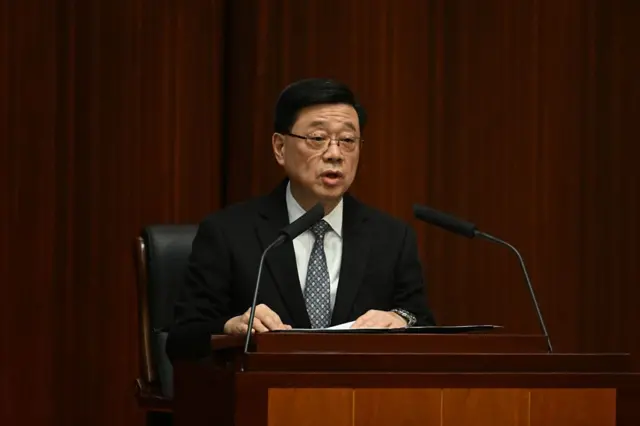 Hong Kong Chief Executive John Lee Ka-chiu sitting at a lectern in a black suit