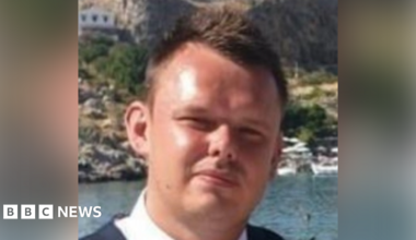 Matthew Adams, who has brown hair, is standing in front of a lake in the sun. In the background there is a cliff, on which a house can be seen, as well as trees and other vegetation. Boats moored on the water can also be seen behind Matthew.
