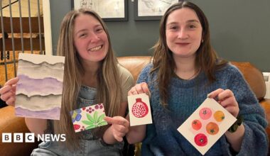 Poppy Barrett (left) and Hannah Broughton (right) are sitting on a brown leather sofa, each holding up small pieces of colourful artwork. The pieces include abstract purple waves, bright green leaves with a pink flower, a red pomegranate, and several circular fruit designs, possibly tomatoes of different colours. Behind them is a dark green wall with framed art.