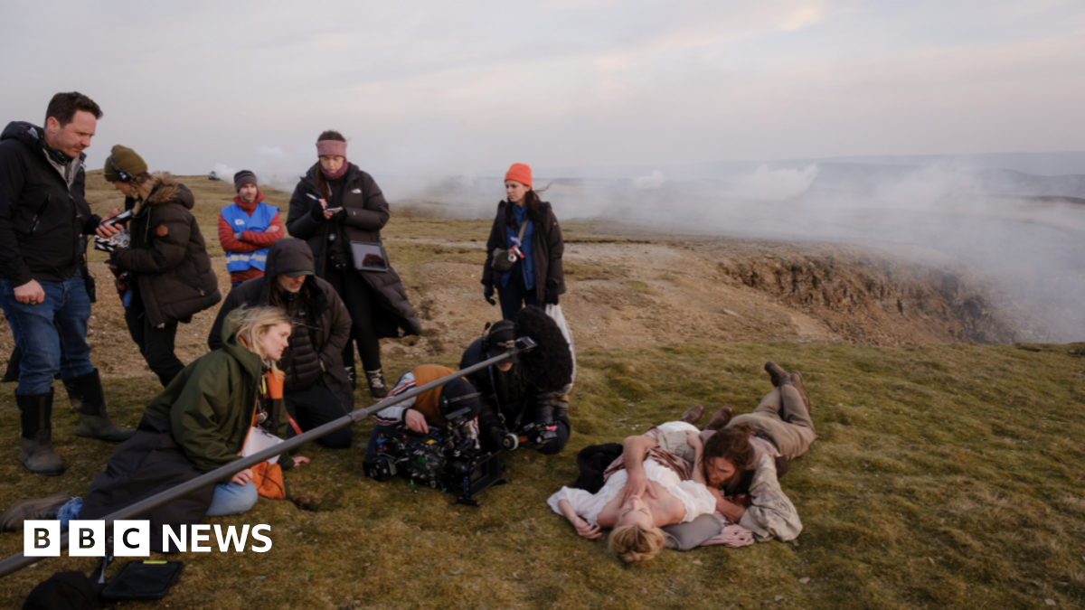 A scene of the 2026 film Wuthering Heights being shot in the Yorkshire Dales. To the left of the image are members of the crew with a boom microphone and camera. To the right Margot Robbie is lying on the ground in costume with Jacob Elordi to her side also in costume