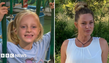 Mary Michelle Devine right in a white dress in a green outdoor area and Theia left in a play area smiling at the camera. She is a blond haired girl wearing a purple t-shirt with a cartoon character on.