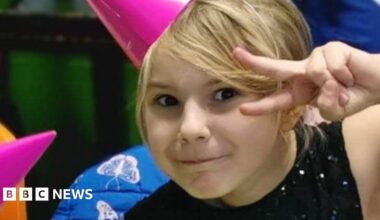 A girl with blonde hair with a side fringe is pulling a cheeky expression at the camera and pulling a peace sign. She is wearing a pink party hat and a black sparkly top.