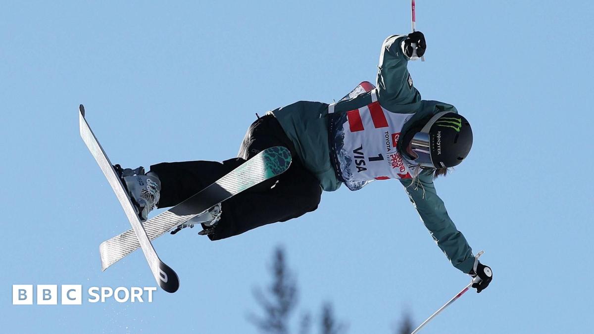 Freestyle skier Zoe Atkin is airborne, with only a blue sky and the tops of trees visible behind her