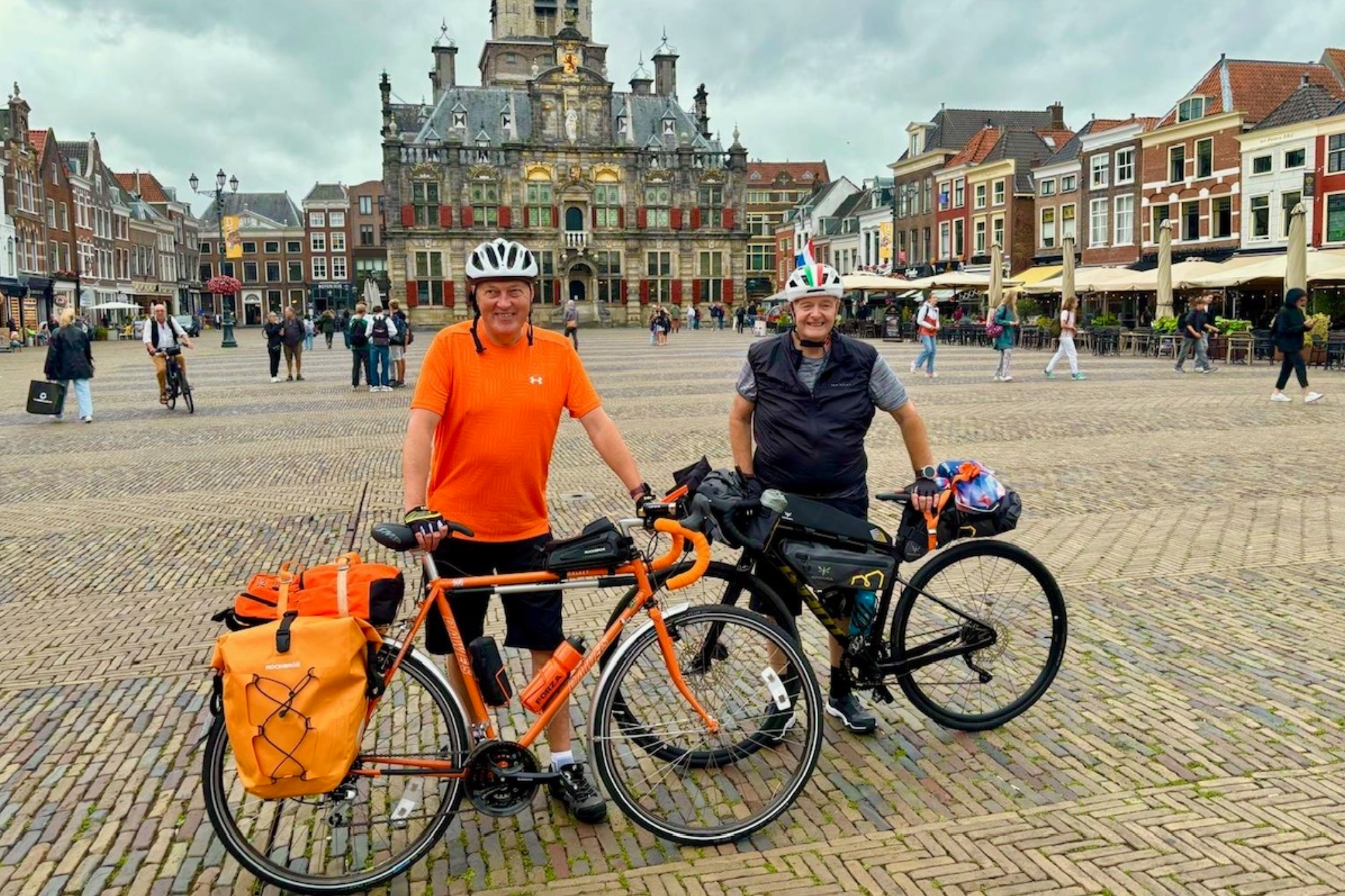 Tony Hall with his orange bike and friend stand smiling at the camara in a town square